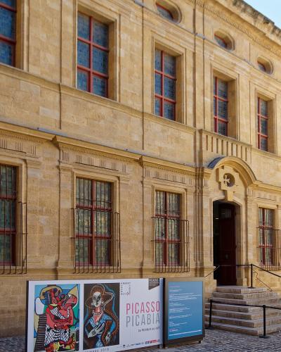 The image shows the exterior of a classical stone building with red-framed windows, barred lower windows, and a central arched entrance; signage in front advertises a "PICASSO PICABIA" exhibition at "GRANET," indicating it is the Musée Granet in A