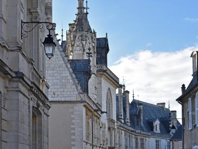A low-angle street view of a historic stone building complex featuring a prominent Gothic-style tower with a spire, set against a blue sky, with cobblestone pavement and bollards in the foreground.