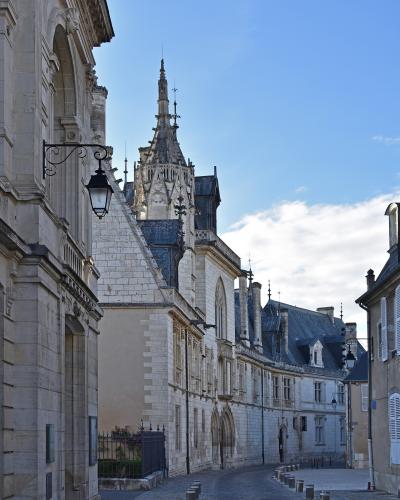 A low-angle street view of a historic stone building complex featuring a prominent Gothic-style tower with a spire, set against a blue sky, with cobblestone pavement and bollards in the foreground.