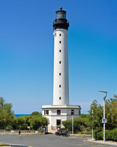 1. A tall, white cylindrical lighthouse with a black lantern room and gallery, standing on a rocky coastal promontory with green vegetation, a paved road in the foreground, and a clear blue sky; no visible signage or text identifying location.