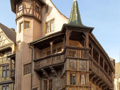 A low-angle photograph of a historic half-timbered building with a prominent corner turret, a conical green-tiled roof, and an elaborate wooden balcony structure, situated on a cobblestone square under a clear blue sky.