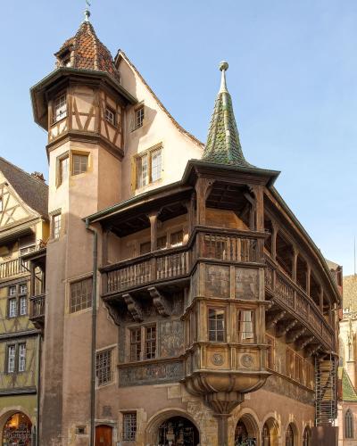 A low-angle photograph of a historic half-timbered building with a prominent corner turret, a conical green-tiled roof, and an elaborate wooden balcony structure, situated on a cobblestone square under a clear blue sky.