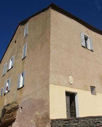 A two-story stone house with a beige stucco facade, white shutters, and a plaque above the entrance, set against a clear blue sky.