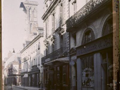 A vintage black-and-white photograph of a cobblestone street in a French city, featuring 19th-century architecture, shopfronts with signage like "A Madame et Bébé" and "TIMBRES ROUGES," and a damaged Gothic tower in the background; the top border rea
