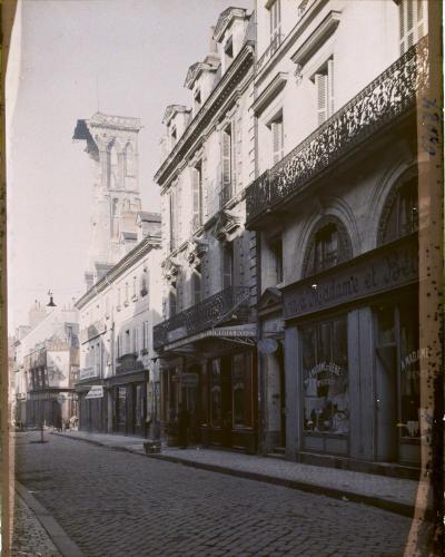 A vintage black-and-white photograph of a cobblestone street in a French city, featuring 19th-century architecture, shopfronts with signage like "A Madame et Bébé" and "TIMBRES ROUGES," and a damaged Gothic tower in the background; the top border rea