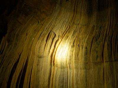 A close-up view of a textured, striated limestone cave wall illuminated by an artificial light source, showing vertical flow patterns and mineral deposits.