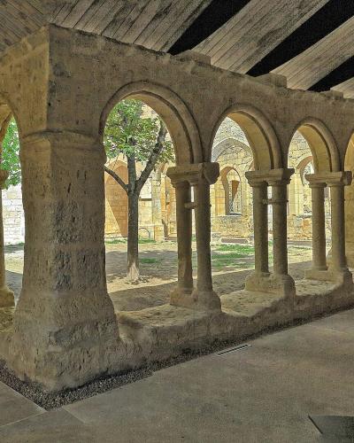 A stone cloister with rounded arches supported by monolithic columns, set within a covered courtyard featuring a wooden-beamed ceiling and a central tree, with modern display elements visible on the floor.