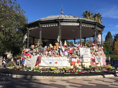 A black metal bandstand (kiosk) in a public park, heavily adorned with stuffed animals, flowers, French flags, and memorial banners including one reading "À NOS ÉTOILES DISPARUES" and another referencing the Nice attack of July 14, 2016.
