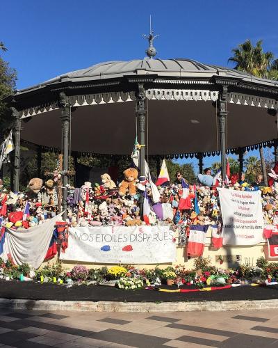 A black metal bandstand (kiosk) in a public park, heavily adorned with stuffed animals, flowers, French flags, and memorial banners including one reading "À NOS ÉTOILES DISPARUES" and another referencing the Nice attack of July 14, 2016.