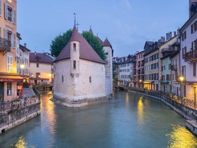 The image shows the Palais de l'Île, a medieval stone building with conical roofs situated on a triangular islet in the Canal du Thiou, flanked by colorful buildings and illuminated streetlamps at dusk.