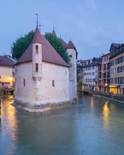 The image shows the Palais de l'Île, a medieval stone building with conical roofs situated on a triangular islet in the Canal du Thiou, flanked by colorful buildings and illuminated streetlamps at dusk.