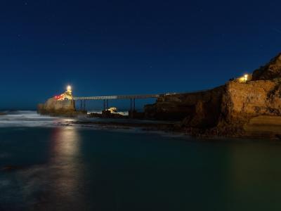 A long-exposure night photograph of a rocky promontory connected to the mainland by a metal truss footbridge, with a brightly lit statue on the rock and red lighting illuminating part of the structure, set against a starry sky over the ocean.