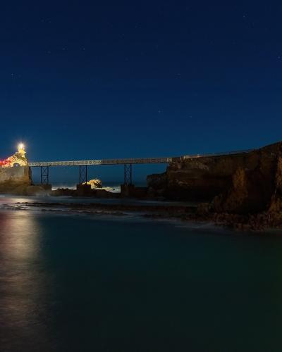 A long-exposure night photograph of a rocky promontory connected to the mainland by a metal truss footbridge, with a brightly lit statue on the rock and red lighting illuminating part of the structure, set against a starry sky over the ocean.