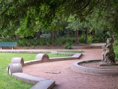 A park scene featuring a stone statue of two figures, curved concrete benches, a green bench, and dense trees.