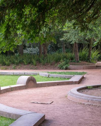 A park scene featuring a stone statue of two figures, curved concrete benches, a green bench, and dense trees.
