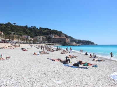 A wide, pebble-strewn beach with numerous sunbathers, turquoise water, and a tree-lined promenade backed by buildings and a green hillside under a clear blue sky.