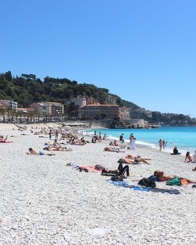 A wide, pebble-strewn beach with numerous sunbathers, turquoise water, and a tree-lined promenade backed by buildings and a green hillside under a clear blue sky.