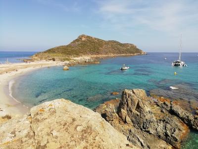 A coastal landscape featuring a rocky headland with sparse vegetation, a sandy beach curving along the shore, clear turquoise water, and boats anchored offshore under a blue sky.
