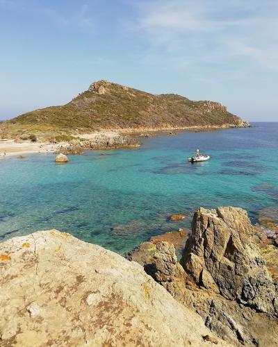 A coastal landscape featuring a rocky headland with sparse vegetation, a sandy beach curving along the shore, clear turquoise water, and boats anchored offshore under a blue sky.