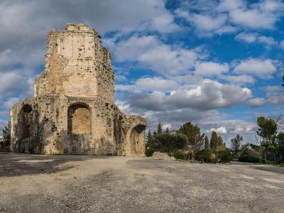 A low-angle photograph of the weathered, octagonal stone ruins of the Tour Magne tower under a partly cloudy blue sky, with gravel ground and surrounding trees.
