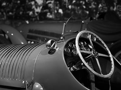 A black-and-white close-up of the cockpit and hood of a vintage racing car, featuring a ribbed metal hood, a large wooden steering wheel, and a cluster of analog gauges.