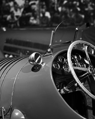 A black-and-white close-up of the cockpit and hood of a vintage racing car, featuring a ribbed metal hood, a large wooden steering wheel, and a cluster of analog gauges.