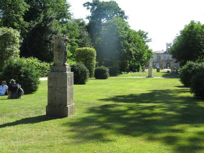 A sunlit park scene featuring a manicured lawn, sculpted hedges, a stone monument in the foreground, and a large classical-style building in the background with people relaxing on the grass.