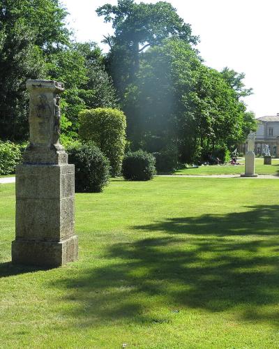 A sunlit park scene featuring a manicured lawn, sculpted hedges, a stone monument in the foreground, and a large classical-style building in the background with people relaxing on the grass.