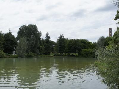 A landscape view of a calm body of water surrounded by dense green trees and vegetation, with a tall brick chimney stack visible in the background on the right side under an overcast sky.