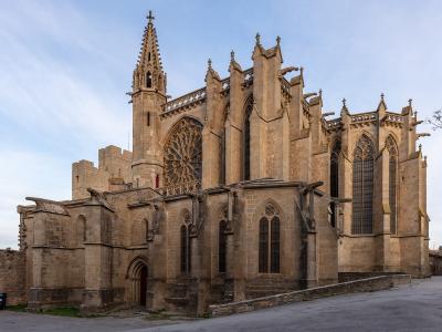 A large Gothic stone church with a prominent rose window, flying buttresses, and a tall spire, situated next to a fortified stone wall under a blue sky.