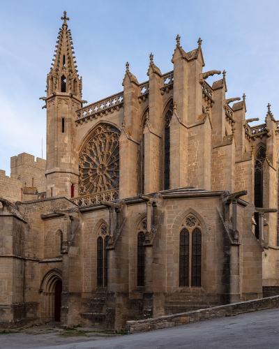 A large Gothic stone church with a prominent rose window, flying buttresses, and a tall spire, situated next to a fortified stone wall under a blue sky.