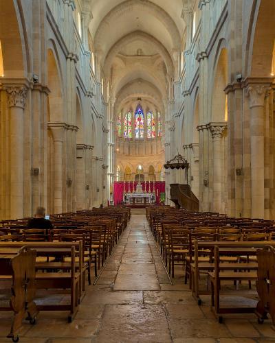 The image shows the interior nave of a large stone church, looking down the central aisle toward the altar, with rows of wooden pews, stone pillars, and a stained-glass window at the far end.