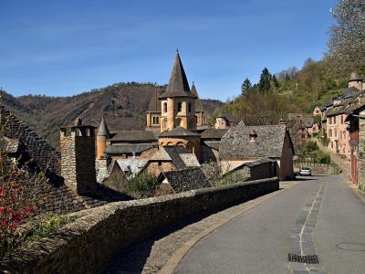 A stone road curves through a hillside village with traditional stone houses and a prominent church featuring a conical spire and Romanesque towers, set against a backdrop of wooded hills under a clear blue sky.