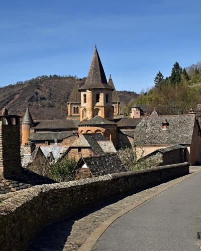 A stone road curves through a hillside village with traditional stone houses and a prominent church featuring a conical spire and Romanesque towers, set against a backdrop of wooded hills under a clear blue sky.
