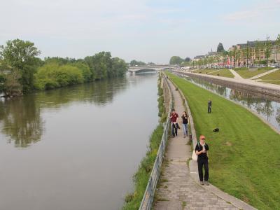The image shows a wide river with a paved walkway and grassy embankment on the right bank, where several people are walking; in the distance, a multi-arch stone bridge spans the river, and buildings line the far bank under a partly cloudy sky.