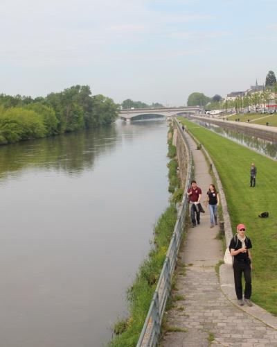 The image shows a wide river with a paved walkway and grassy embankment on the right bank, where several people are walking; in the distance, a multi-arch stone bridge spans the river, and buildings line the far bank under a partly cloudy sky.