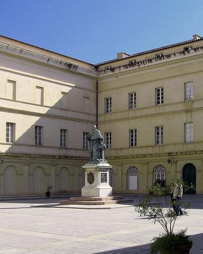 A sunlit courtyard of a large, pale yellow neoclassical building with arched ground-floor arcades and a central statue on a pedestal.