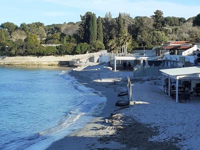 A coastal beach scene showing a sandy shore with calm blue water, backed by a stone seawall, dense pine and cypress trees, and white buildings with terracotta roofs on the right.