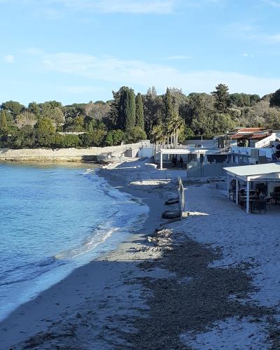 A coastal beach scene showing a sandy shore with calm blue water, backed by a stone seawall, dense pine and cypress trees, and white buildings with terracotta roofs on the right.