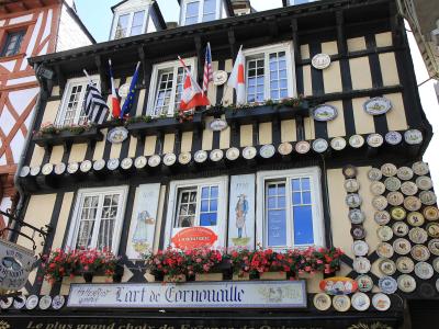 A half-timbered building facade decorated with numerous ceramic plates, flags, and flower boxes, featuring signage for "Faïencerie de Quimper," "Henriot Quimper," and "L'art de Cornouaille."