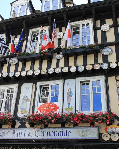 A half-timbered building facade decorated with numerous ceramic plates, flags, and flower boxes, featuring signage for "Faïencerie de Quimper," "Henriot Quimper," and "L'art de Cornouaille."