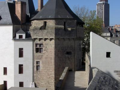 The image shows a stone medieval tower with a conical slate roof, part of a larger castle complex featuring white-walled buildings and brick chimneys, with a modern high-rise building visible in the background under a clear blue sky.