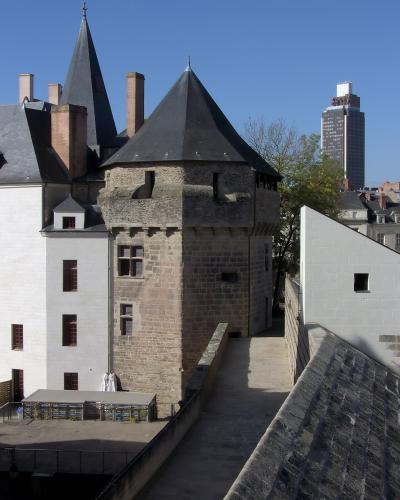The image shows a stone medieval tower with a conical slate roof, part of a larger castle complex featuring white-walled buildings and brick chimneys, with a modern high-rise building visible in the background under a clear blue sky.