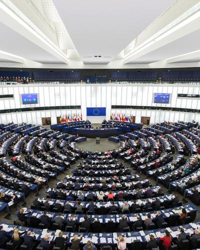 1. The image shows a large, semi-circular hemicycle chamber filled with seated delegates at desks, featuring a prominent European Union flag at the front, multiple national flags, and tiered public viewing galleries; signage includes digital screens 