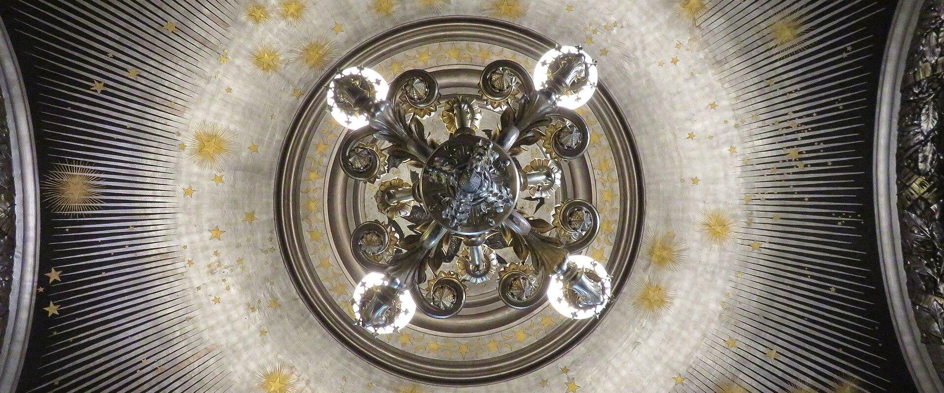 A close-up, upward-looking view of an ornate ceiling featuring a central chandelier with multiple lit bulbs, surrounded by a circular pattern of radiating lines and golden star motifs on a dark background.