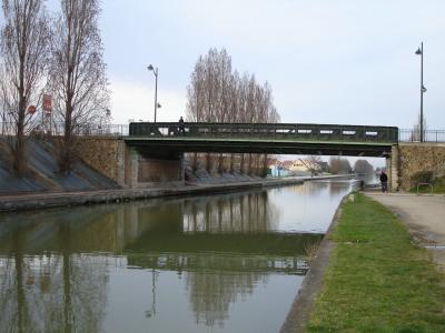 A green metal truss bridge crossing a canal with stone abutments, bare trees, and a pedestrian path alongside the water.