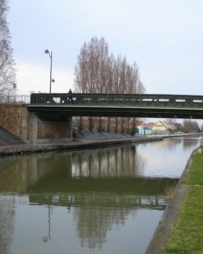 A green metal truss bridge crossing a canal with stone abutments, bare trees, and a pedestrian path alongside the water.
