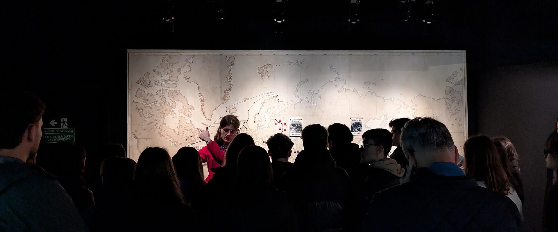 A group of people are gathered in a dimly lit indoor space, listening to a guide in a red jacket who is pointing at a large illuminated map on the wall; a green sign on the left reads "SORTIE DE SECOURS" (Emergency Exit).
