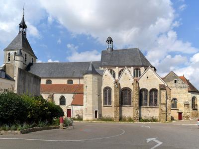 The image shows a stone church with a prominent octagonal bell tower on the left, a main nave with a gabled roof and a small lantern tower, and a side chapel with large arched windows, situated next to a paved road with cars and a building with de