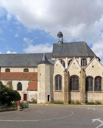 The image shows a stone church with a prominent octagonal bell tower on the left, a main nave with a gabled roof and a small lantern tower, and a side chapel with large arched windows, situated next to a paved road with cars and a building with de